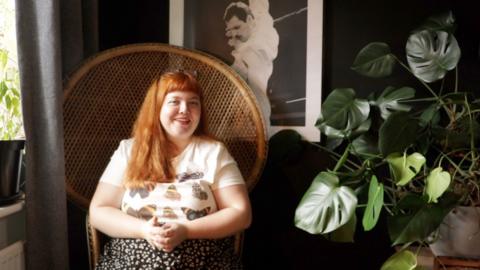 A woman with long red hair is smiling and sitting in a brown armchair. There is a plant to her left and a poster of Freddie Mercury behind her.