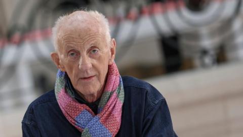 Paul Costelloe has receding grey hair. He is wearing a bright pink, blue and green scarf and a blue jacket.
