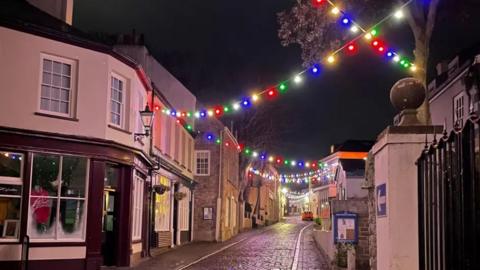 A cobblestone street with Victorian buildings at night decorated with colourful lights.