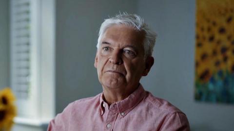 Phillip Schofield looking sombre, in a pink shirt in a room with sunflowers plus a sunflower picture, filming for episode two of Channel 5's Cast Away.