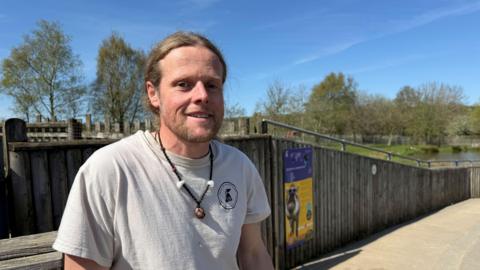 Mike Wilson, a man wearing a light grey shirt and necklace, is standing outdoors next to a wooden fence. Behind him is an animal enclosure with trees and pools of water. There is a blue and yellow sign on the fence.