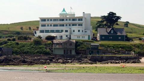 Burgh Island Hotel.  White with a green top of it. A smaller dark building sits to the right and a smaller building again further down the hill in front of the hotel is also visible.  The hill continues up behind the hotel.