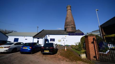 The outside of Moorcroft Pottery in Burslem, featuring a large chimney on top of a white building. Cars can be seen outside.