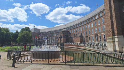 The City Hall building on a sunny day. There are a few clouds in the sky and a water fountain with a stream of water surrounding it.