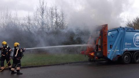 Firefighters spray water onto the back of a bin lorry, which is on fire