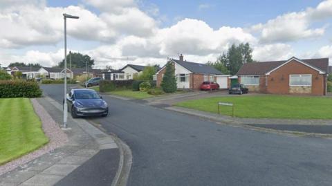 A general view of the brown-orange bricked bungalows, green lawned gardens and grey road of Rivington Drive in Bury