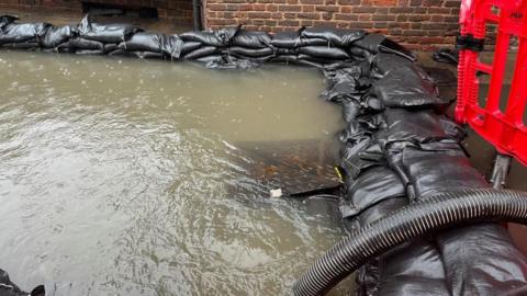 Flooding in Chalfont St Peter. It shows a number of black sand bags and piping.