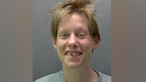 Erin McNeilly is seen in a police custody image the woman with short brown hair, smiling. She is wearing a grey round-necked T-shirt and one of her front teeth is missing.