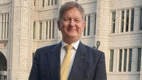 Alex Nicoll, man smiling in suit, shirt, and yellow tie, in front of granite council headquarters building