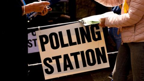 Two people facing one another. We cannot see their heads. The one of the right is wearing a pink coat and is holding a clipboard. In between them is a white sign with the words POLLING STATION written in black.