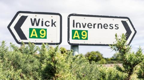 Two road signs on the A9 showing the directions to Wick and Inverness. The signs are white with black arrows pointing the way. In yellow in a green box is the word "A9". In black letters are the names "Wick" and "Inverness".