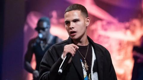 Slowthai performing holding a microphone with a light orange and purple background