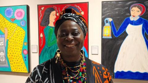 The photo shows Chinwe Russell smiling at the camera. She is wearing a patterned jacket and bright beaded necklace. She is standing in front of three bright coloured portraits, all of women. 