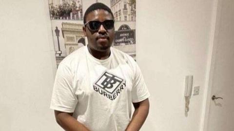 A man with short dark hair and wearing dark glasses is wearing a white Burberry T-shirt and standing in front of a poster on a white wall.