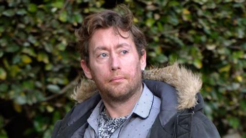 A man in his late 30s with wavy brown hair, a blue parka and a blue shirt outside the High Court in Glasgow. There are bushes in the background of the image and he looks to the right of where the camera is.