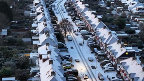 Aerial view of snow-covered rooftops of houses lining a road, also covered in snow