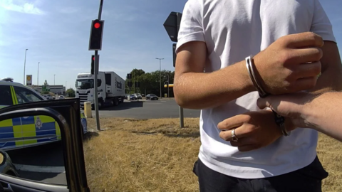 A man in a white t-shirt with handcuffs on next to a police car