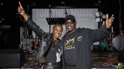 Beverley Knight, wearing a black leather jacket and T-shirt, is standing next to a man in a black hoodie and T-shirt that says "Diversity Music" on it. They are smiling at the camera with one arm each held up in the air. Behind them are several microphones and guitars and a screen saying "Together Music" on it.