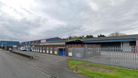 A long brick building with lots of white windows on an industrial estate. There is a metal fence on one side and a yard behind it.