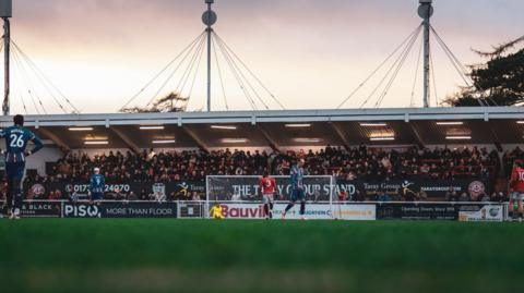Players from Chatham Town, in a red kit, and Carlisle United, in a blue kit, play on the pitch in front of a stand at the Bauvill Stadium.