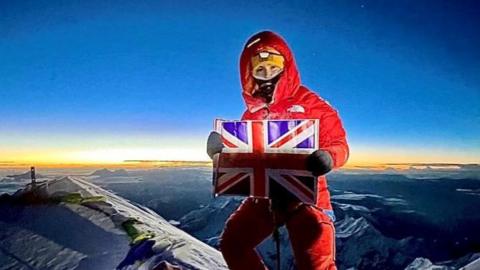 Adriana Brownlee stands at the peak of a mountain in a red snowsuit, yellow hat and black mask, holding a Union Jack flag, with a sunset behind her