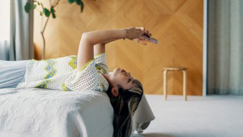 Woman lays on bed looking at her phone in a modern home with a house plant in the background.
stock image