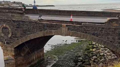 Collapsed section of Banff Bridge. A section of brickwork has falled to the area below. The sea is in the backgroundm with a single orange and white cone on the bridge. 