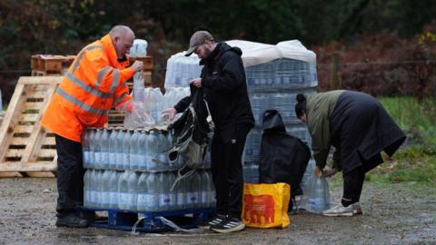 A man in high visibility jacket standing in a car park with a lot of bottles of water 