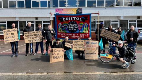 A picket line outside the school. Thirteen people are holding placards that read "transparent pay = workers who stay" and "give us a break". "Safety at work", reads another", "all about the kids, not just the marketing". The national education union banner is in the middle, reading "education is a right not a privilege". 