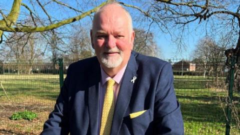 Police and Crime Commissioner Jonathan Evison wearing a blue suit with a yellow tie, standing in a field with a barbed wire fence and trees in the background