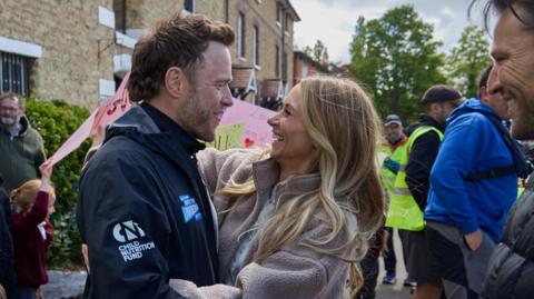 Olly Murs and his wife Amelia look at each other and embrace. There is a crowd around him, some of which are holding signs. Murs is wearing a black sports jacket and Amelia is wearing a brown fleece and has long blonde hair. 