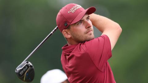Jack Whaley, in red golf shirt and cap, tees off and watches his shot