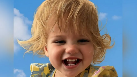 Jayla-Jean Mclaren, a toddler with blonde hair wearing a brightly coloured dress, smiles at the camera. 