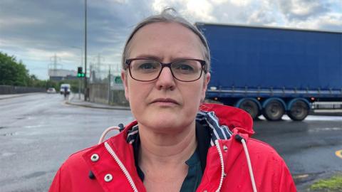 Lizzi Collinge standing at a crossroads by the port in a red coat. She has fair hair tied back and is wearing glasses. In the background a large blue lorry passes to the right