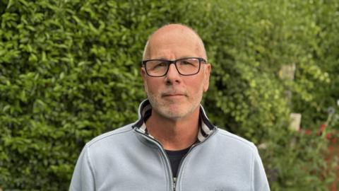 Phil Dankert looks directly into the camera while standing in front of a large hedge which covers the whole background. He is bald with white stubble. He is wearing black glasses, a navy blue t-shirt and a light blue fleece.