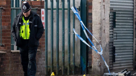 A police officer at the scene where Gavin Parry, 31, was found dead at the scene on Western Road in Birmingham. There is old loose blue police tape on dark green metal railings blowing in the wind.
