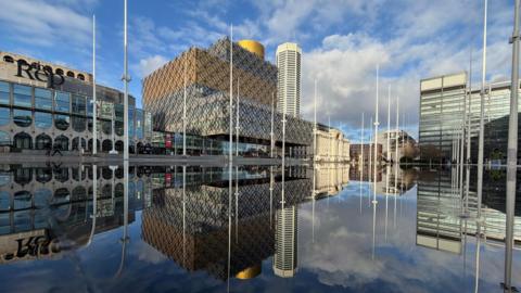 A cityscape under a partially cloudy sky, reflected in a still sheen of water