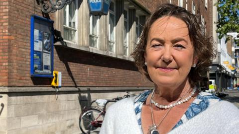 Joanna Brittan is looking at the camera and smiling. She has a short dark bob and is wearing a pearl necklace and earrings. She wears a white fluffy cardigan and is pictured from the shoulders up. She is standing outside a police station.