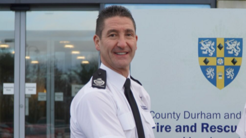Fire officer Keith Carruthers stands in front of a white office building with automatic opening doors. He has short brown/grey hair and wears a fire officer uniform, which is white with black badges on his shoulders and a black tie.