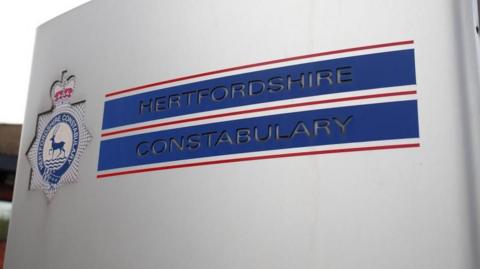 Hertfordshire Constabulary's steel sign, with writing etched in blue bands with parallel red lines in between. The force's crest, with a stag in the middle, is to the left of the sign.