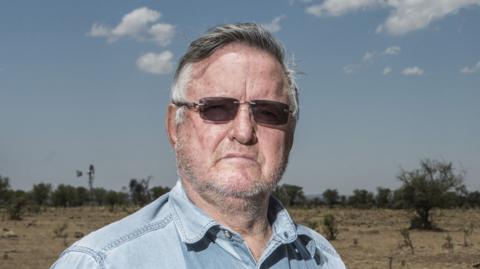 John Hume looks into the camera, wearing rimless sunglasses and a blue denim shirt