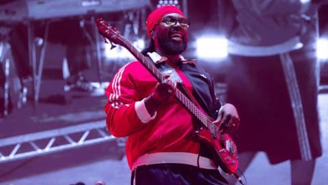 Black man with beard and glasses smiling. He is wearing a red hat, red tracksuit top and playing a red guitar.