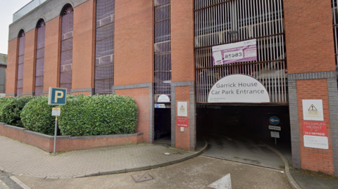 A brick building that is bring used as a multi-storey car park. Outside the building is a blue sign with a white P inside it and a large hedge row. A white arched sign also reads Garrick House Car Park Entrance. 