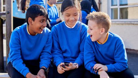 Three children sat in a playground holding tv micro:bits. They are wearing blue sweatshirts and are smiling.