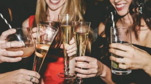 Library shot of a group of women holding glasses with straws and liquid in them. Two of the women's faces are visible - one has long blond hair and the other has long dark hair. One is wearing a red dress and the other has a black dress. They are enjoying themselves.