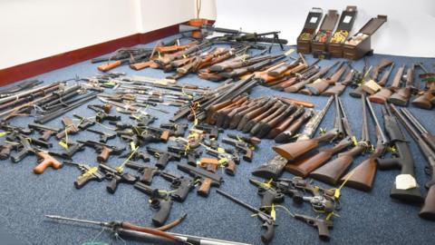A wide shot of dozens of different guns and knives laid out on a blue-grey carpet in a room with white walls.