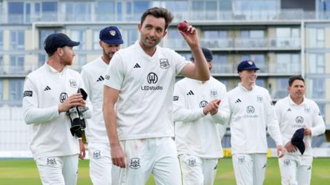 Glos seamer Matt Taylor holds the ball aloft in front of his team-mates after taking five wickets against Northants