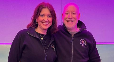 A woman with dark hair hugging a bald man man in a radio studio with a purple backdrop 
