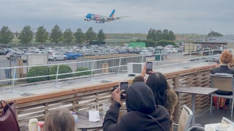 A colourful airplane descends towards an airport while onlookers take photos from a nearby observation area.