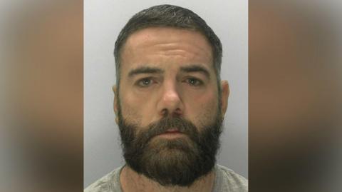 Police headshot of Robert Hall. He has a full beard and short brown hair, brown eyes and is wearing a grey t-shirt.